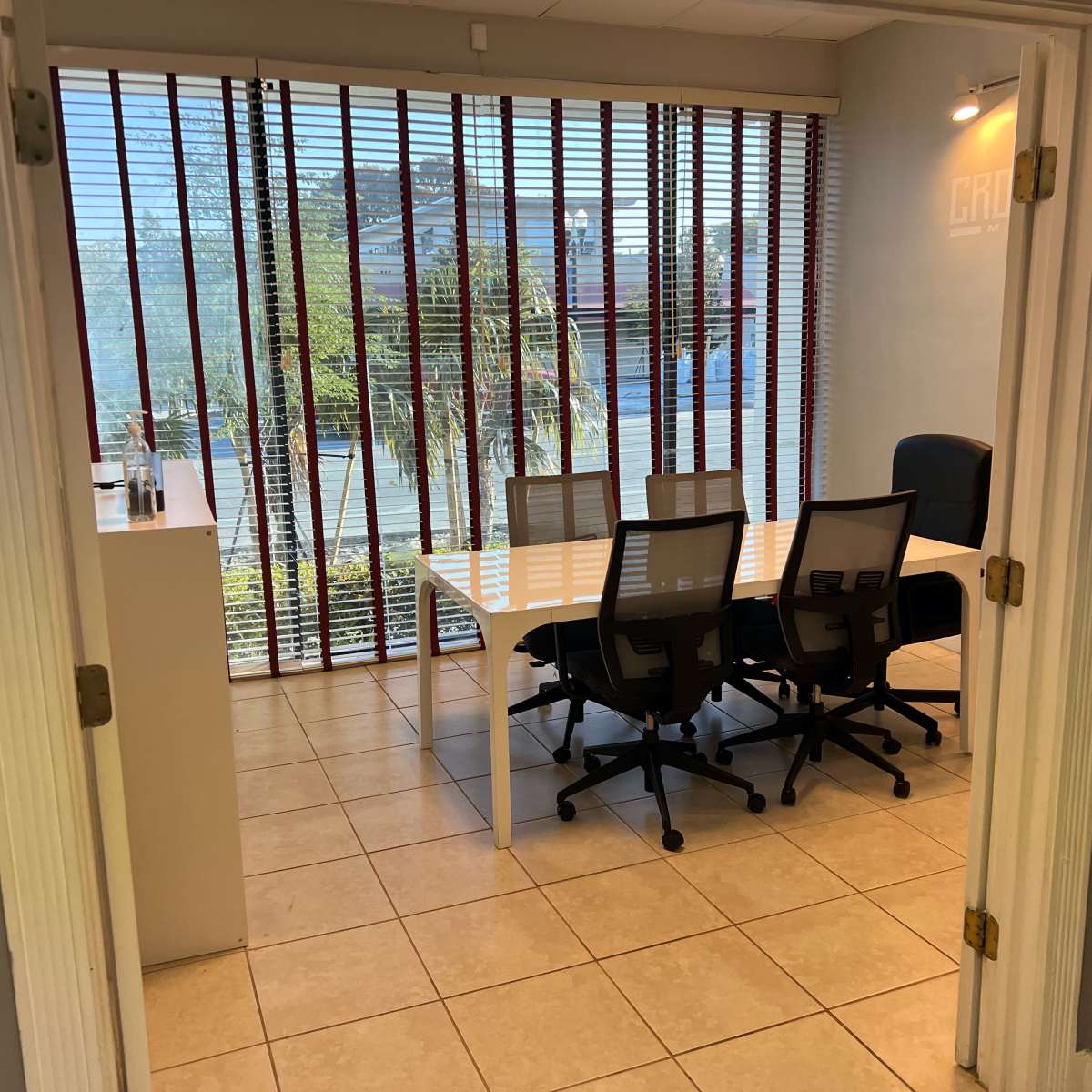 A modern office space with a conference table and chairs, illuminated by natural light through vertical blinds.