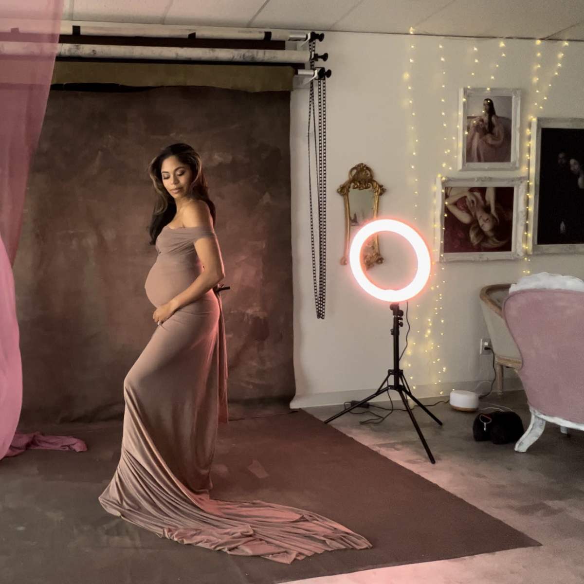 A pregnant woman in a form-fitting gown poses in a photography studio with fabric drapes and soft lighting.