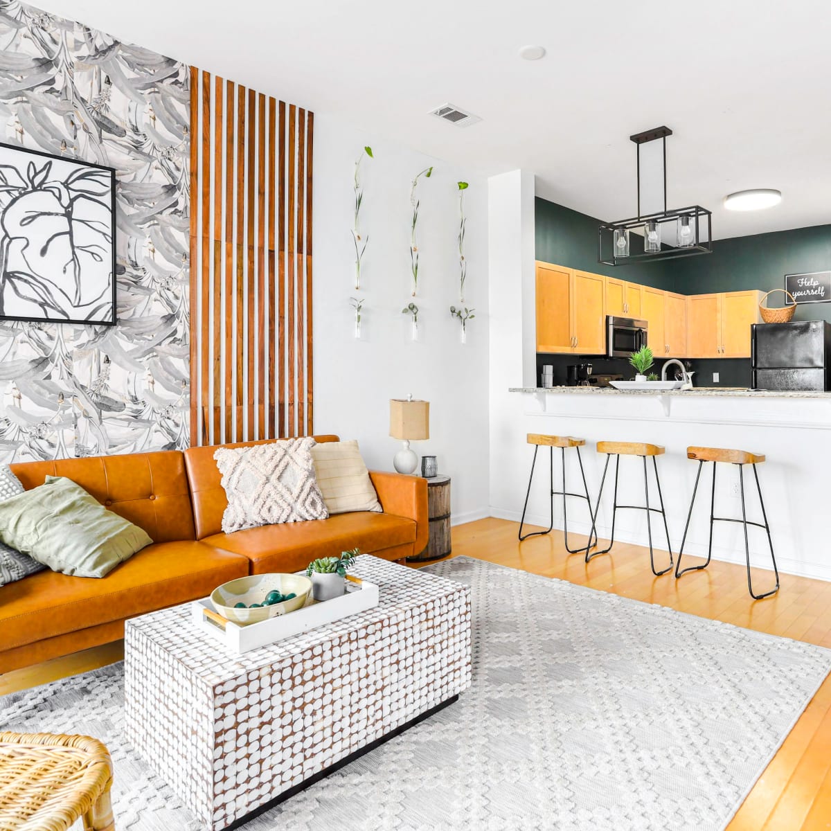 A modern living space featuring a brown leather sofa, a patterned area rug, a coffee table, and a kitchen area with wooden cabinets and bar stools.