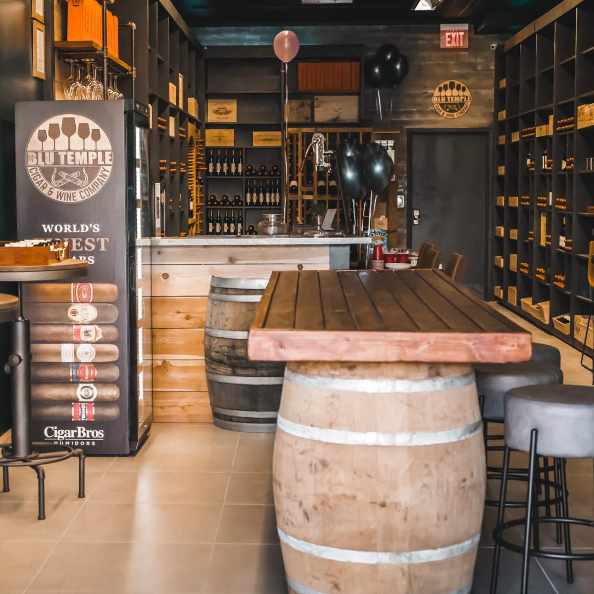 The interior of a bar featuring wooden tables, barrels, and shelves lined with bottles.