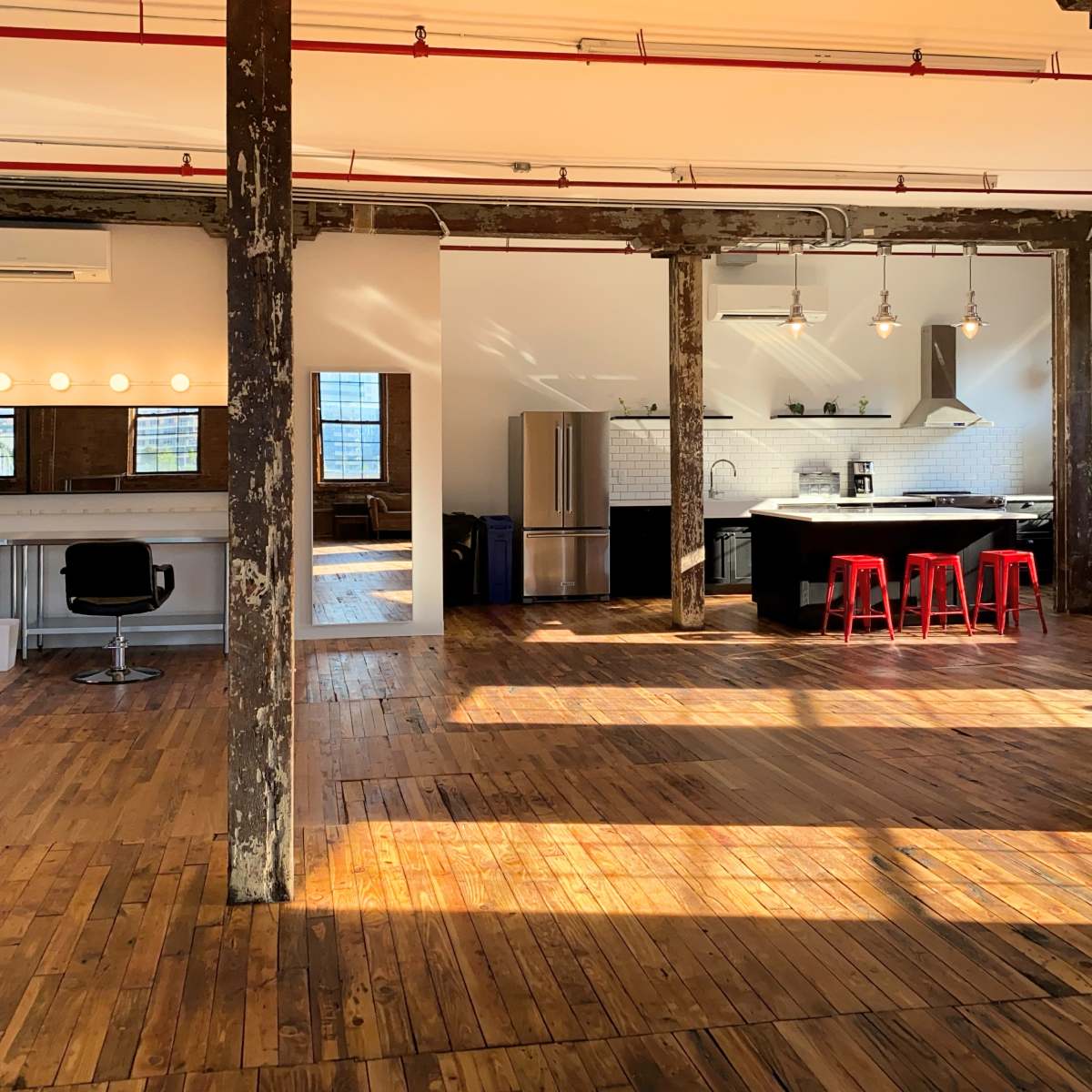 A spacious, industrial-style interior with wooden floors, exposed pillars, and a kitchen area featuring stainless steel appliances and red bar stools.