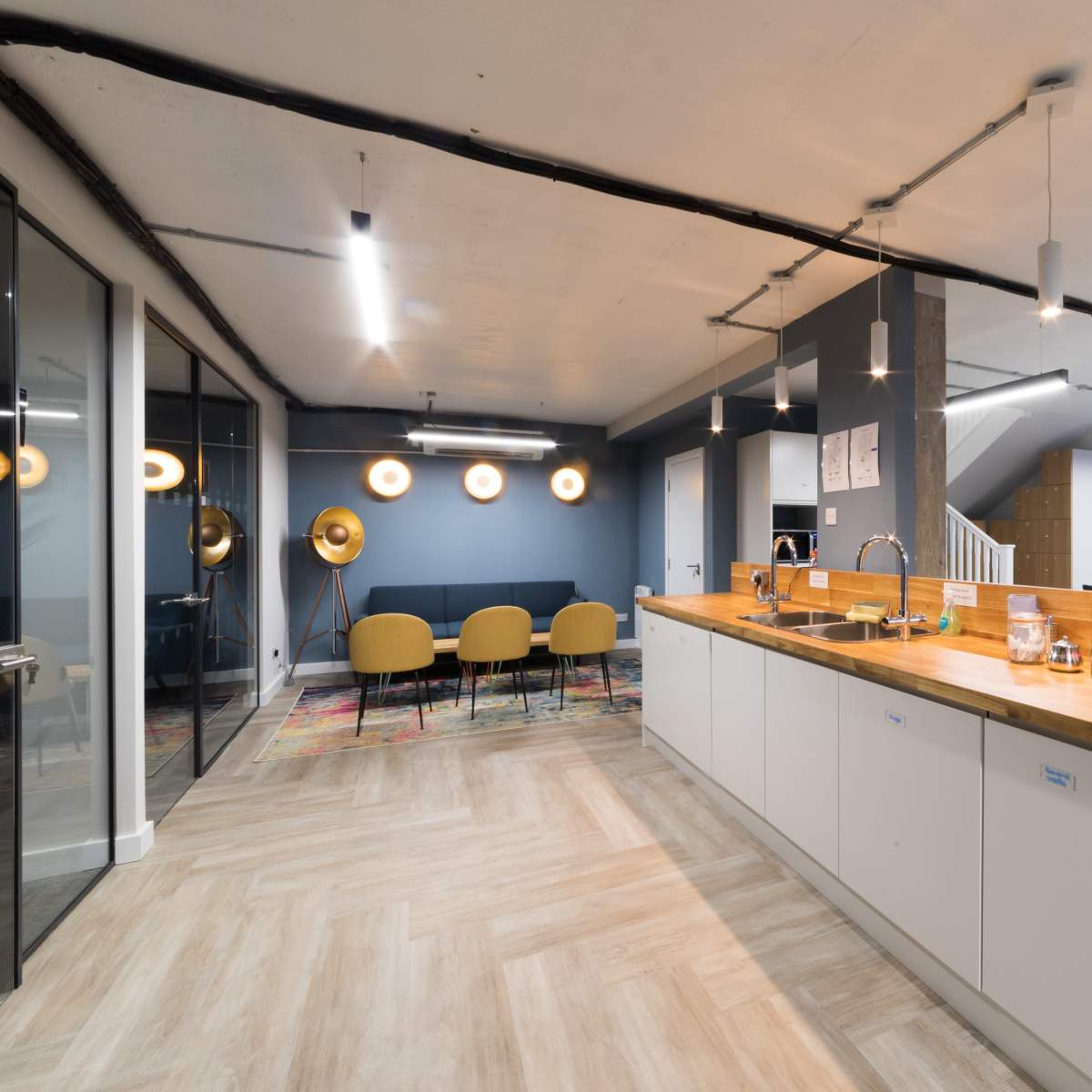 A modern office space with a kitchen area featuring a wooden countertop, a seating area with yellow chairs, and workstations in the background.