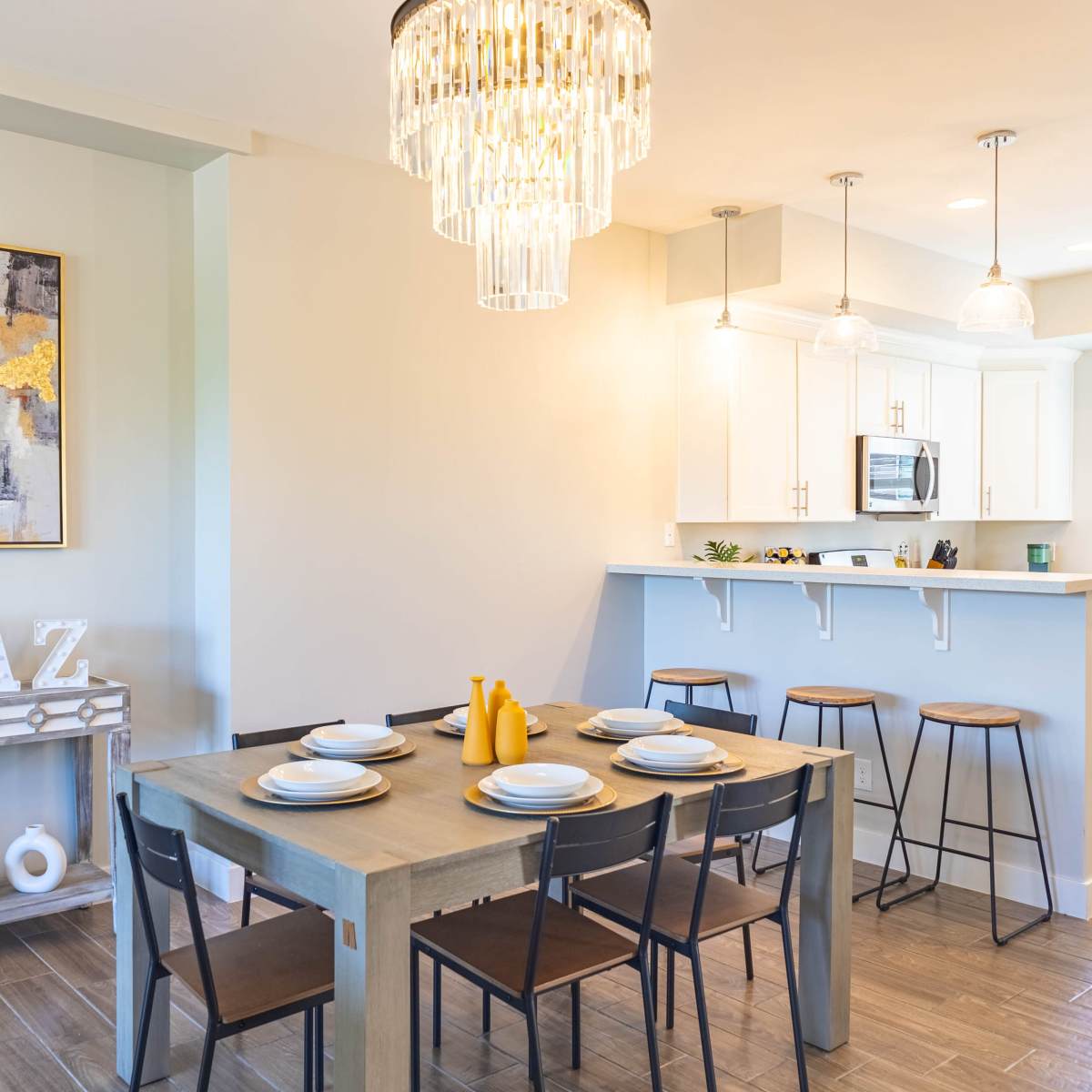 A modern dining area features a rectangular table set for six, a stylish chandelier overhead, and a well-lit kitchen in the background.
