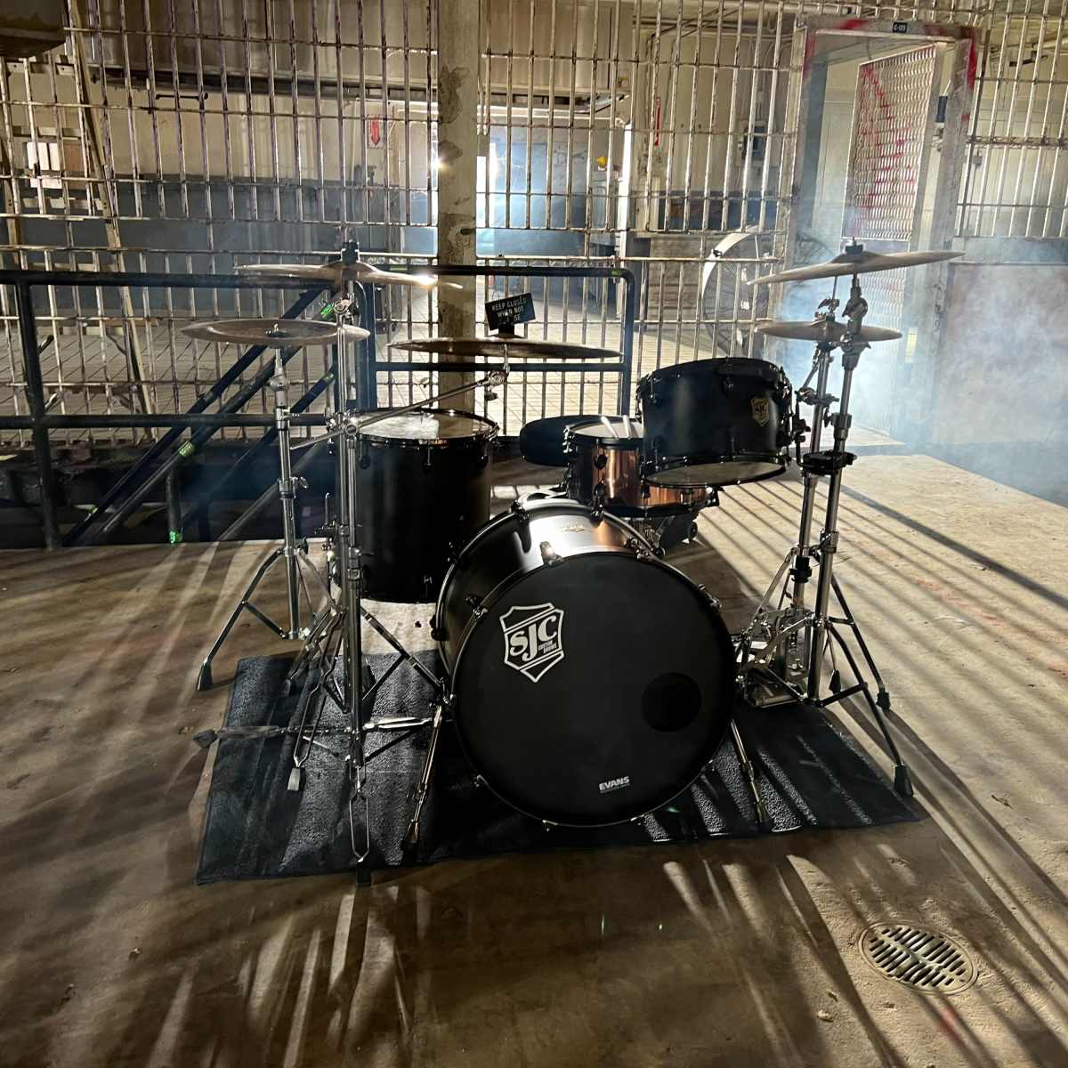 A drum set sits in a dimly lit, industrial space with metal bars in the background and smoke lingering in the air.