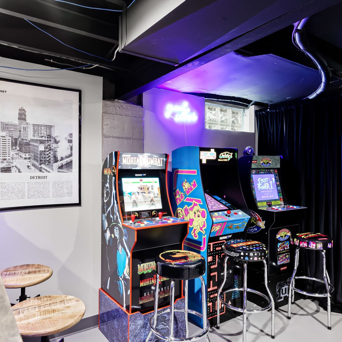 A gaming corner in a basement featuring arcade machines, a framed black and white cityscape photograph, and bar stools.
