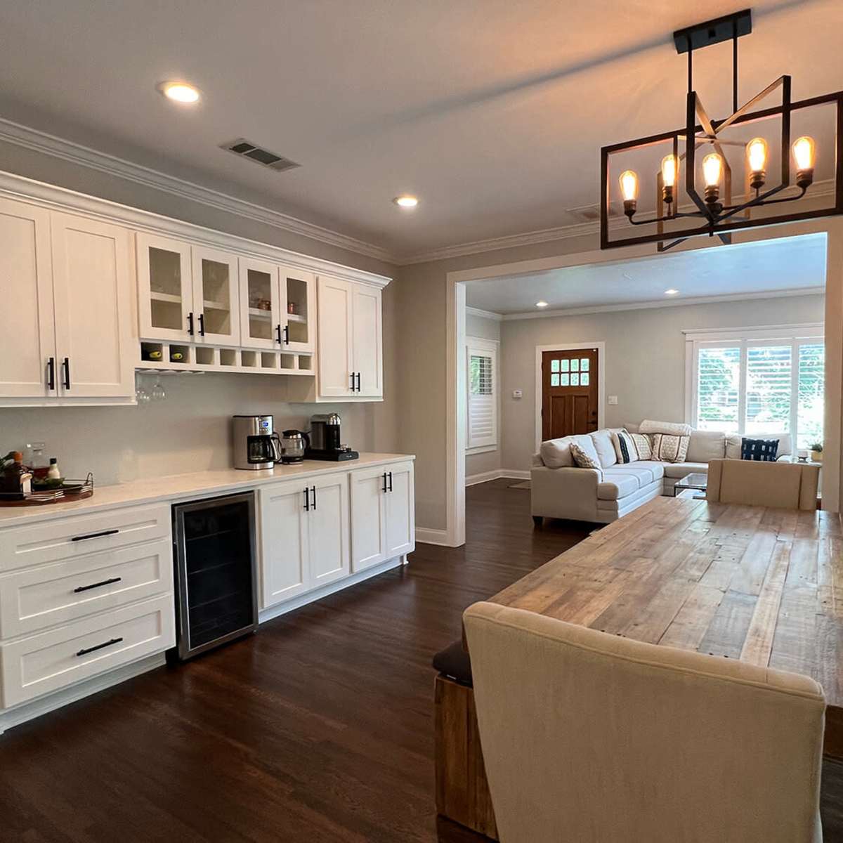 A modern kitchen and dining area featuring white cabinetry, a wooden dining table, and a living room visible in the background.