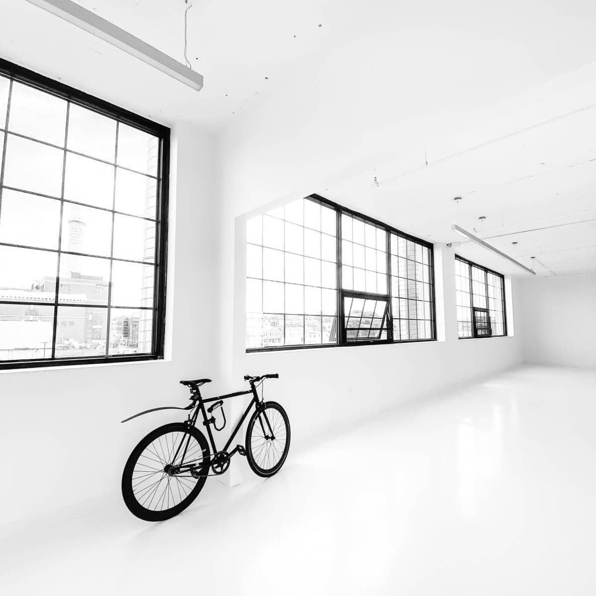 A bicycle is positioned next to a row of large windows in a bright, minimalist room with white walls and a polished floor.