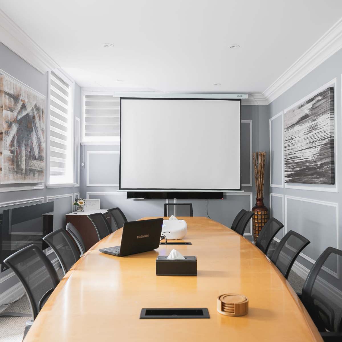 A modern conference room features a long wooden table, black chairs, and a wall-mounted projector screen, with artwork and windows along the walls.