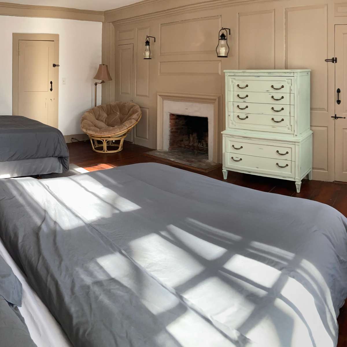 A bedroom with two large beds, a light green dresser, a chair, and a fireplace, illuminated by sunlight casting shadows across the wooden floor.