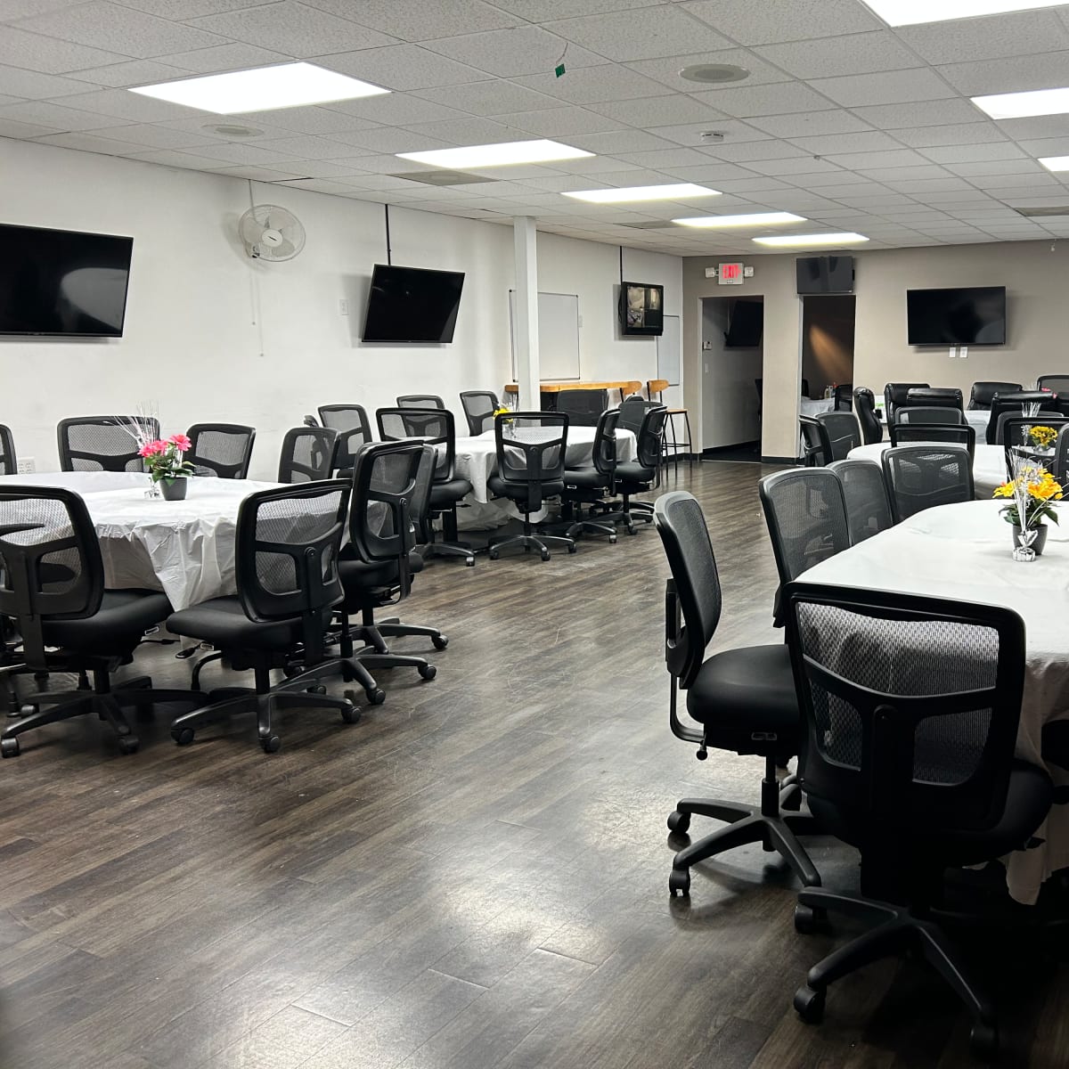 The room features multiple round tables set with white tablecloths and black office chairs, along with television screens mounted on the walls.