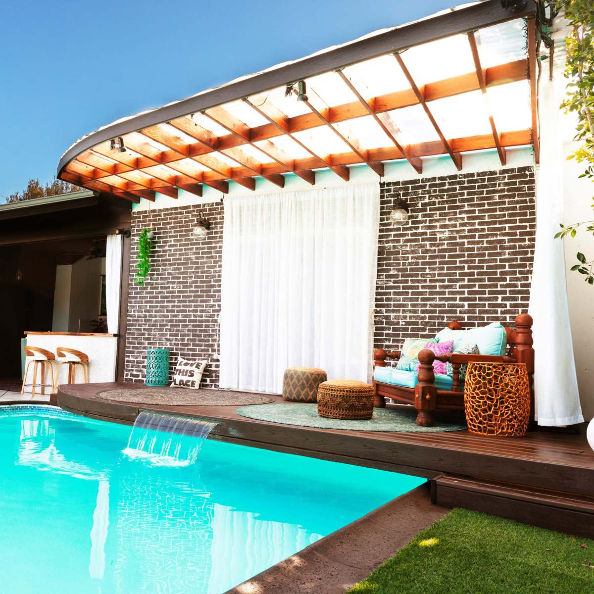 A modern backyard features a swimming pool with a water feature, surrounded by a patio and a brick wall, with comfortable seating and greenery.