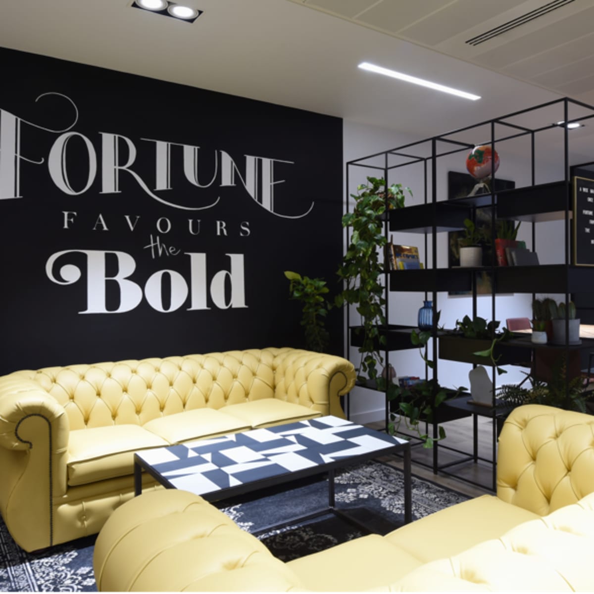 A modern office lounge area featuring yellow chesterfield sofas, a black and white patterned coffee table, and a wall with the phrase "Fortune Favours the Bold" in stylish lettering.