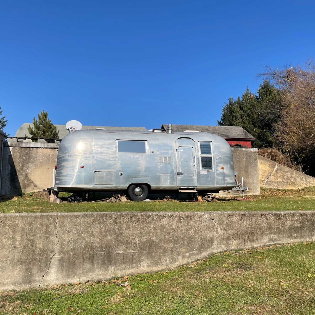 A vintage Airstream trailer is parked on a grassy area next to a concrete wall under a clear blue sky.