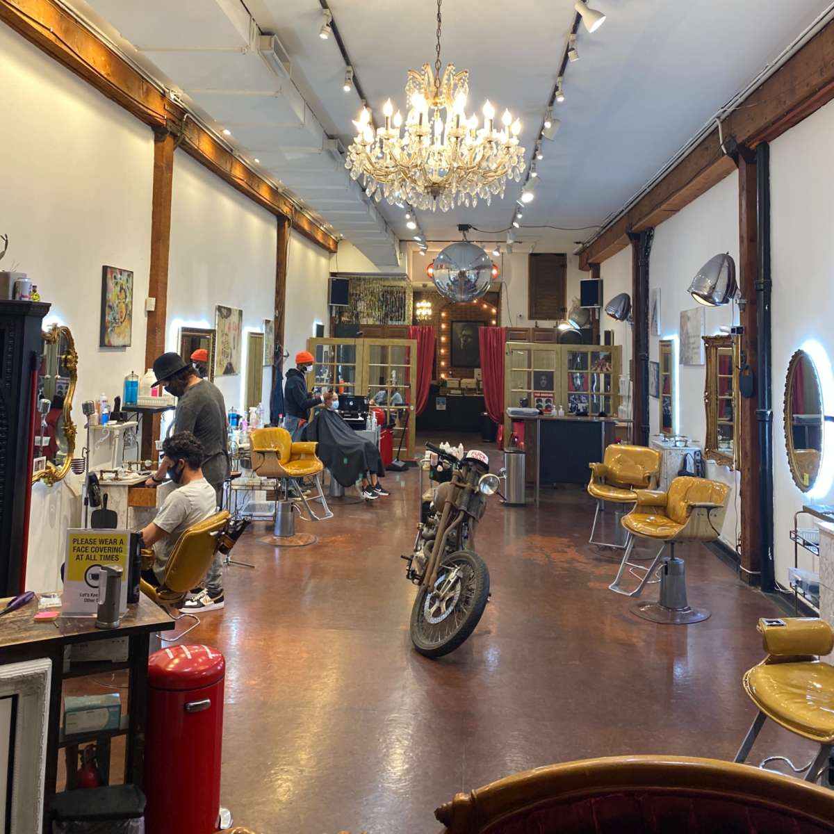 The interior of a barbershop features vintage yellow chairs, a chandelier, and a motorcycle positioned in the center of the floor.