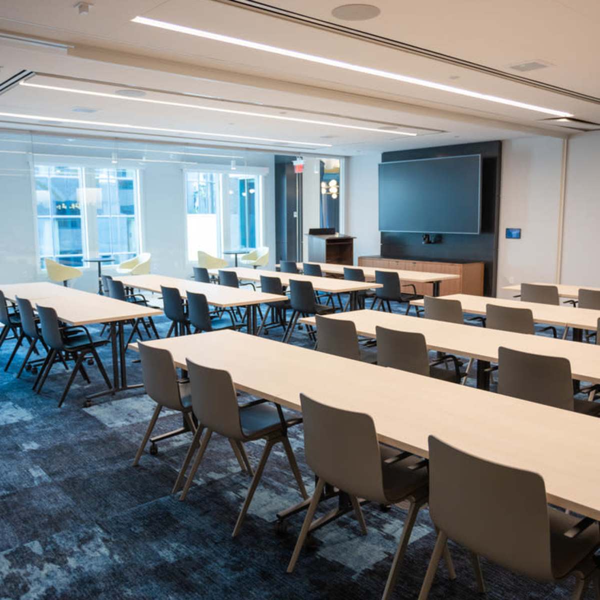 A spacious conference room features several long tables arranged in rows, with chairs facing a large screen at one end.