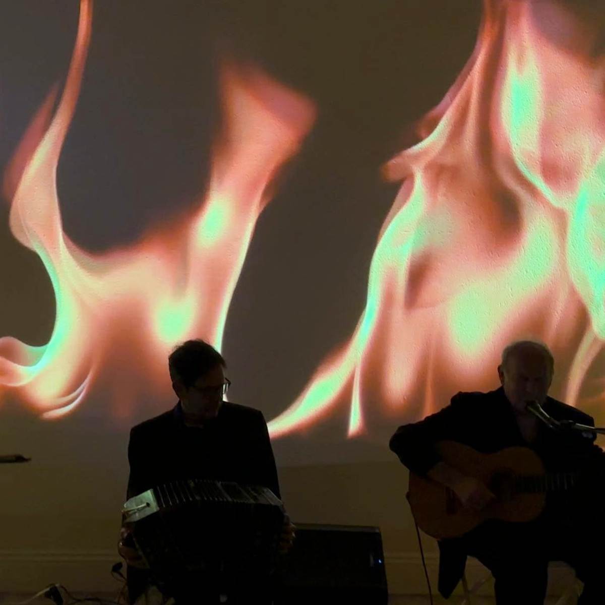 A musician plays a double bass in front of a backdrop featuring animated flames, while two other musicians accompany him on guitar and a different instrument.