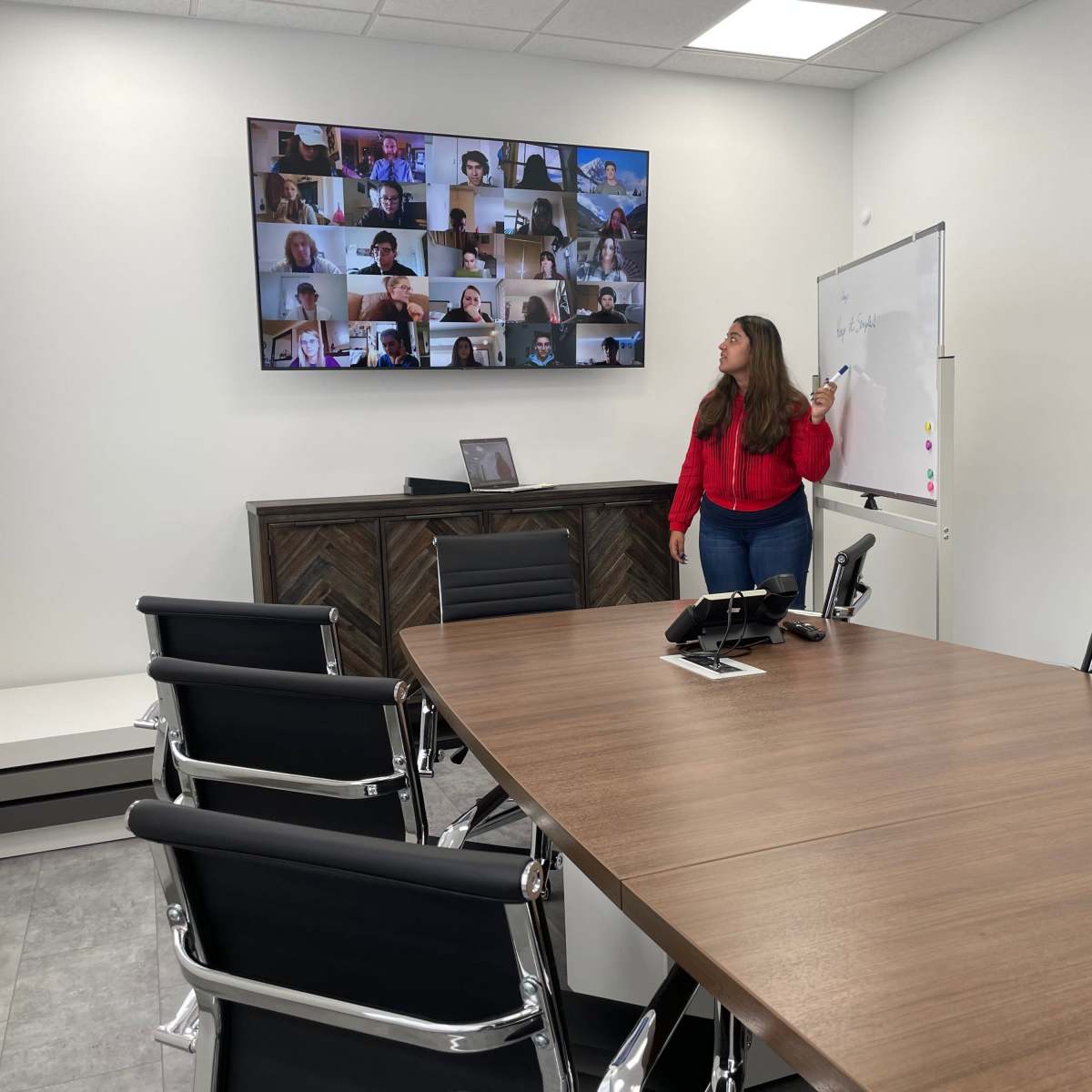 A woman stands near a conference table, pointing at a whiteboard while a large screen displays a video conference with multiple participants.