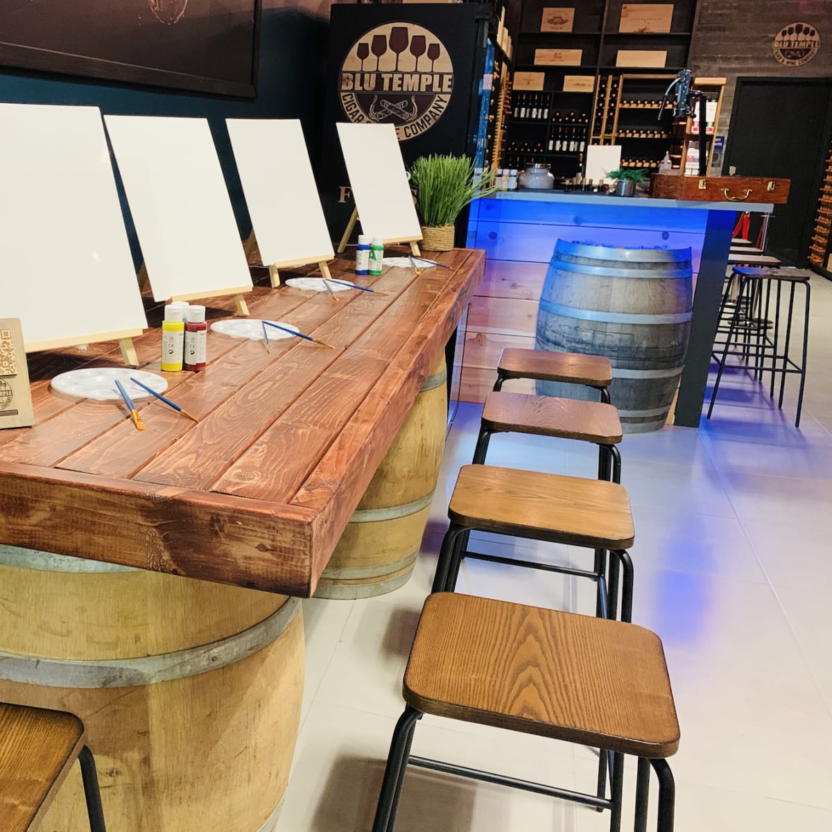 A wooden bar counter made from barrels features blank canvases and art supplies, with wine bottles and glasses positioned nearby.
