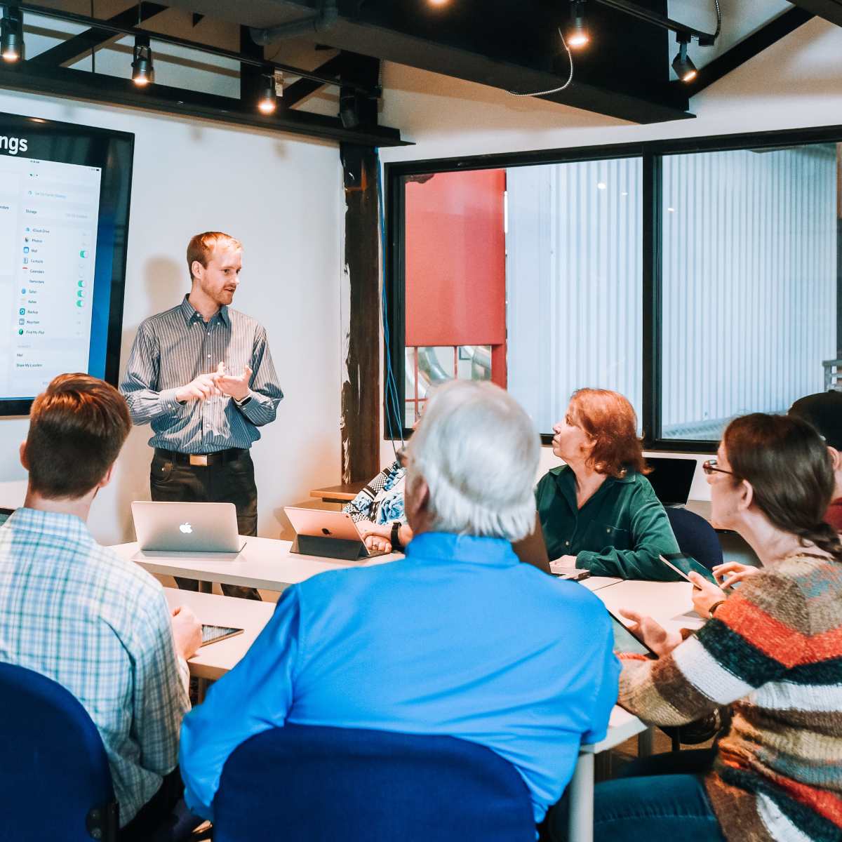 A presenter stands in front of a group of seated individuals in a conference room, engaging them while a large screen displays iCloud device settings.