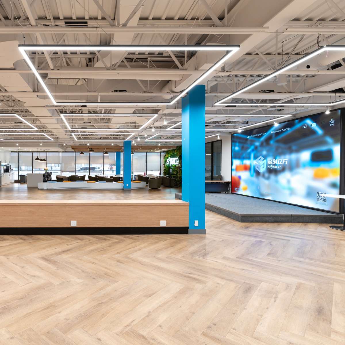 A modern office space featuring wooden flooring, bright lighting, and a reception area with a large digital display in the background.