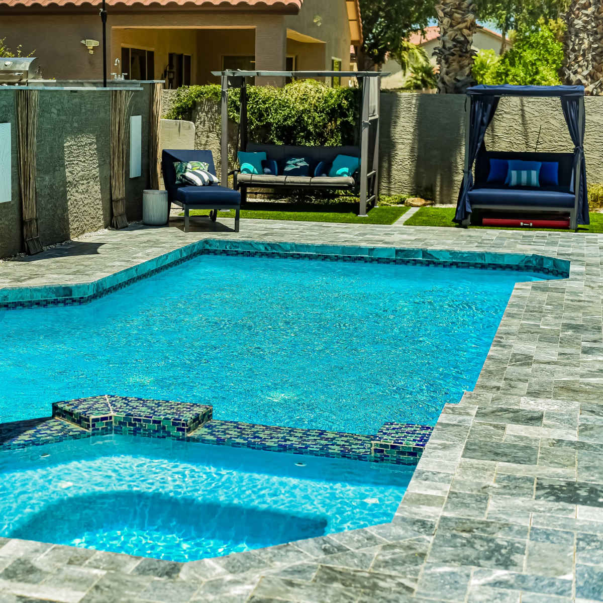A rectangular swimming pool surrounded by gray stone patio tiles and lounge chairs in a backyard setting.