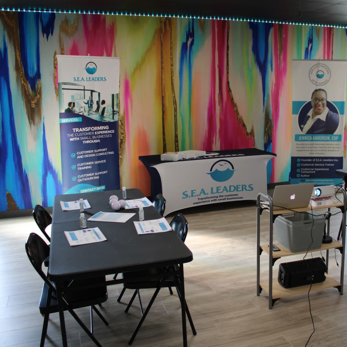 A training room setup with tables, chairs, promotional banners, and a projector against a colorful mural.