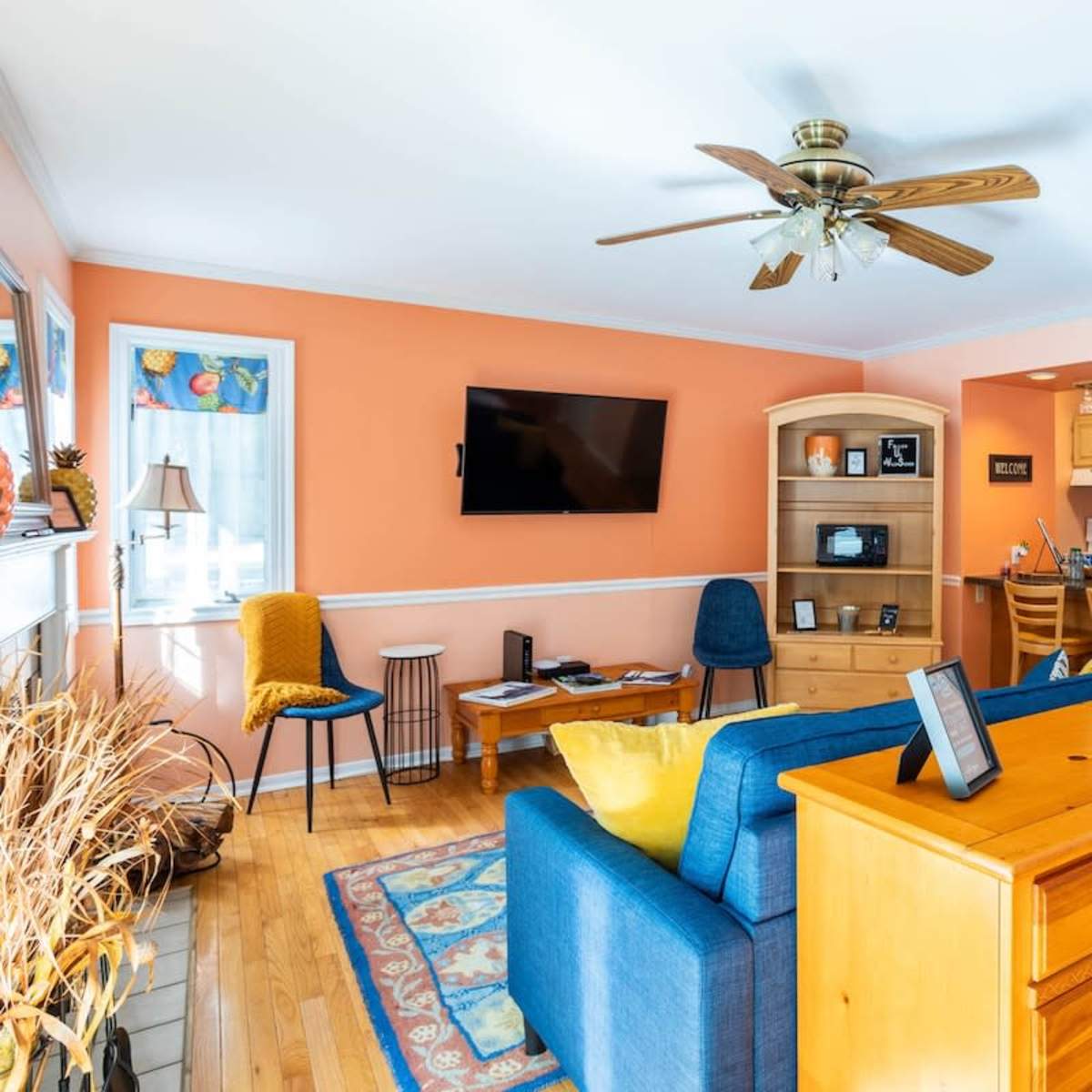 A bright living room with an orange wall, a blue sofa, wooden furniture, and a kitchen area visible in the background.