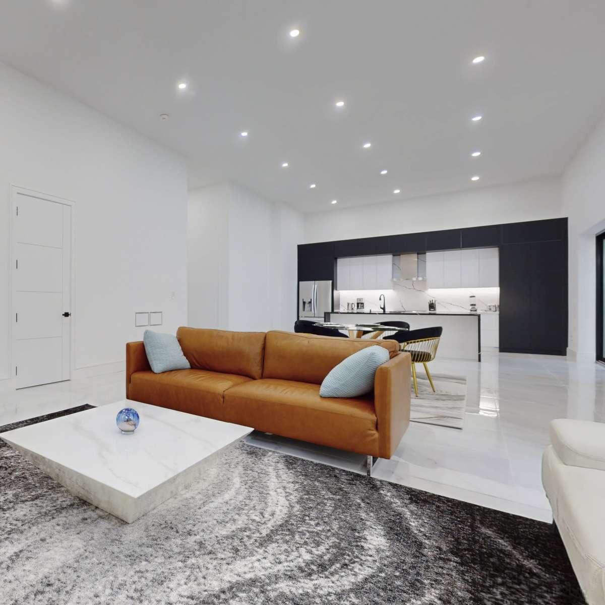 A modern living room with a brown sofa, a marble coffee table, and a staircase leading to an upper level, with a kitchen visible in the background.