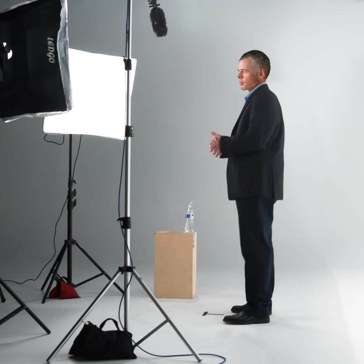 A man in a suit stands beside a microphone and a box on a plain white backdrop, with lighting equipment set up around him.