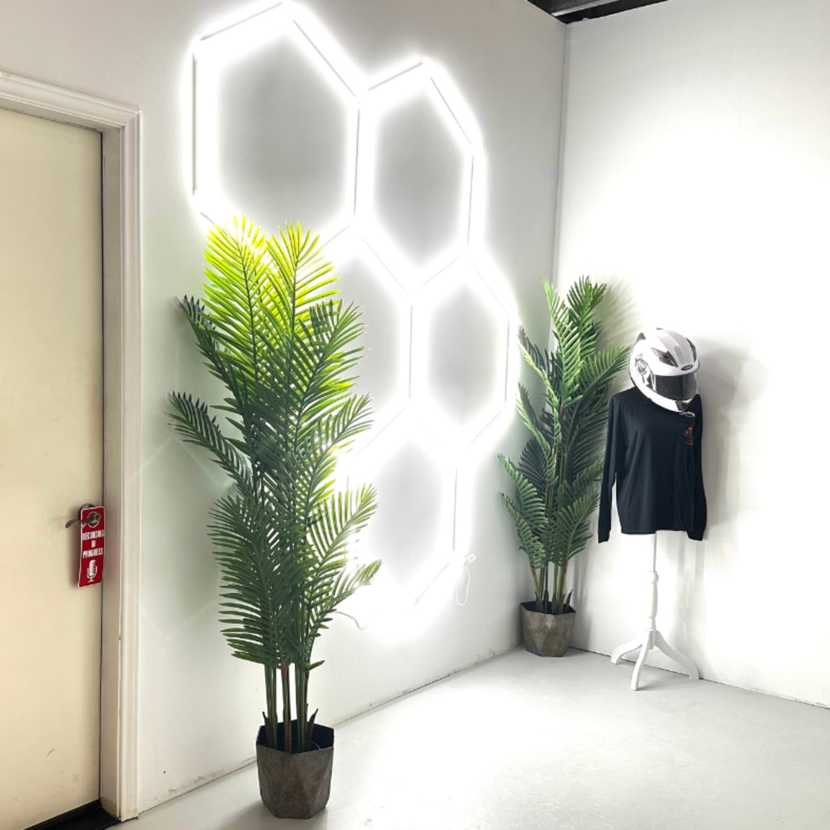 A corner of a room featuring two potted palm plants, a mannequin wearing a black shirt and a helmet, and a hexagonal neon light installation on the wall.