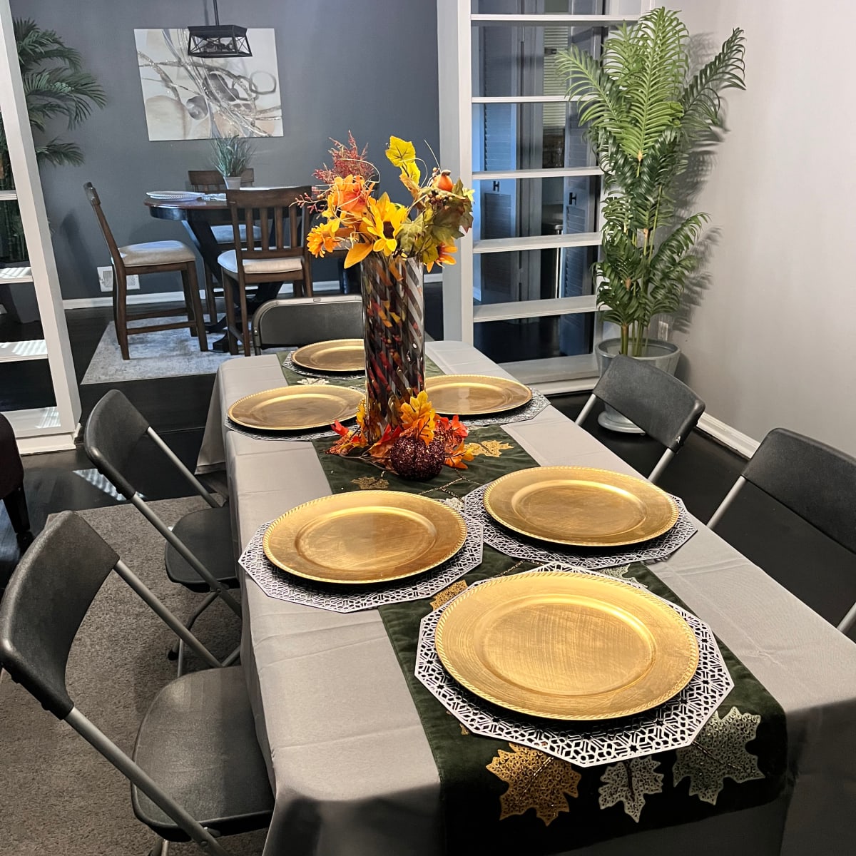 A formal dining table is set with gold plates and a centerpiece of autumn flowers, while covered by a gray tablecloth and surrounded by folding chairs.