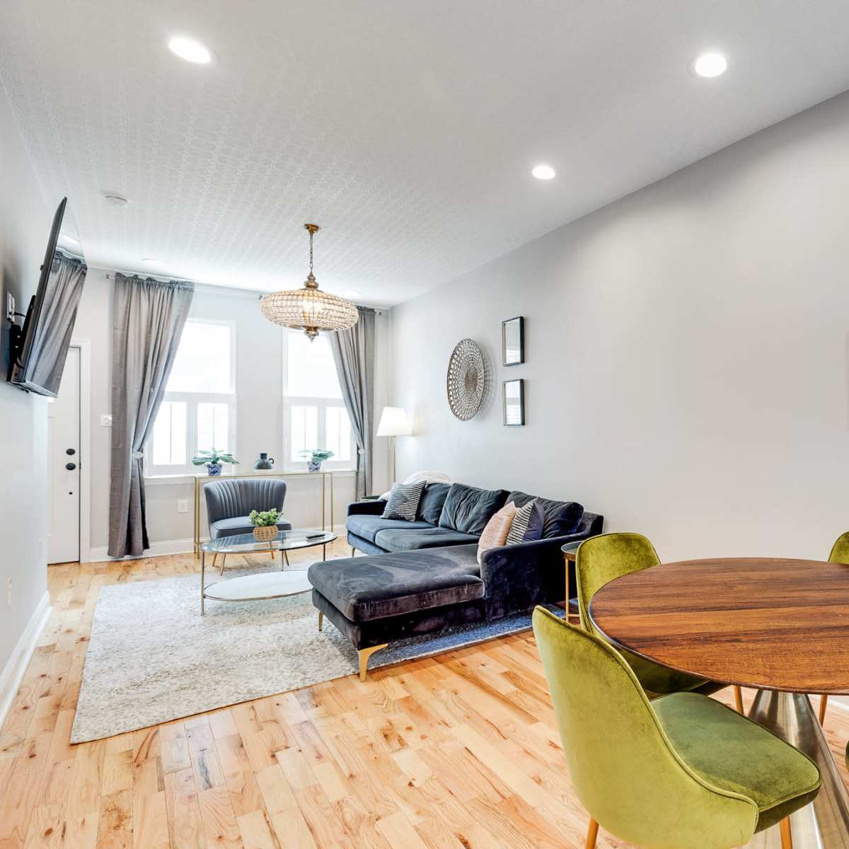 A modern living space featuring a dark sofa, a round wooden dining table with four green chairs, and a potted plant against a light gray wall.