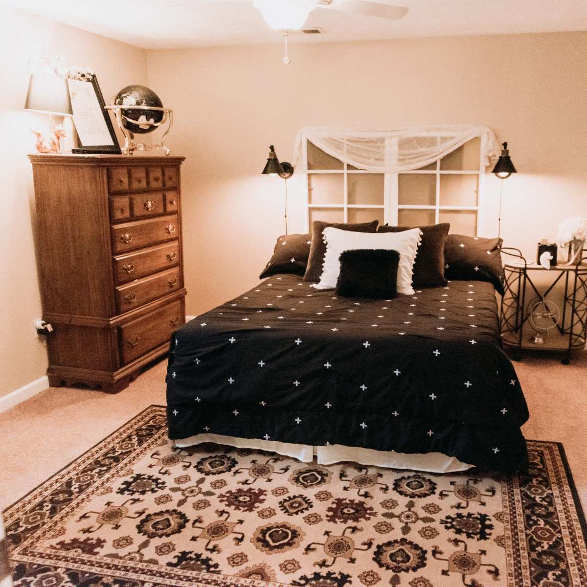 A bedroom features a bed with a dark patterned comforter, a wooden dresser, and a decorative rug on the carpeted floor.