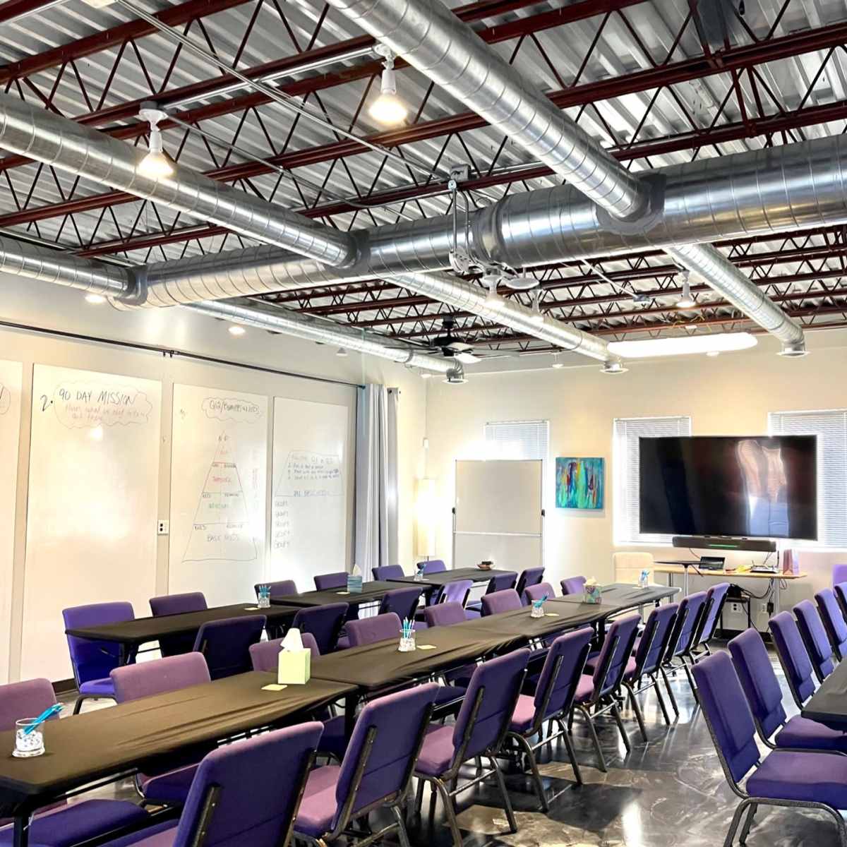 A spacious conference room is set up with multiple tables and purple chairs for a meeting, featuring whiteboards, a large screen, and visible ceiling ducts.
