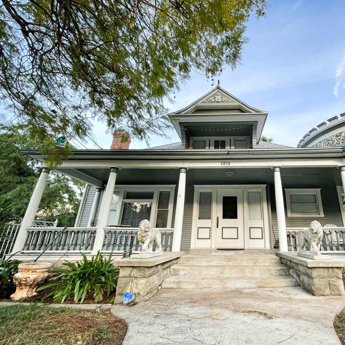 A large, historic house with a wraparound porch, ornate architectural details, and lush greenery in the front yard.