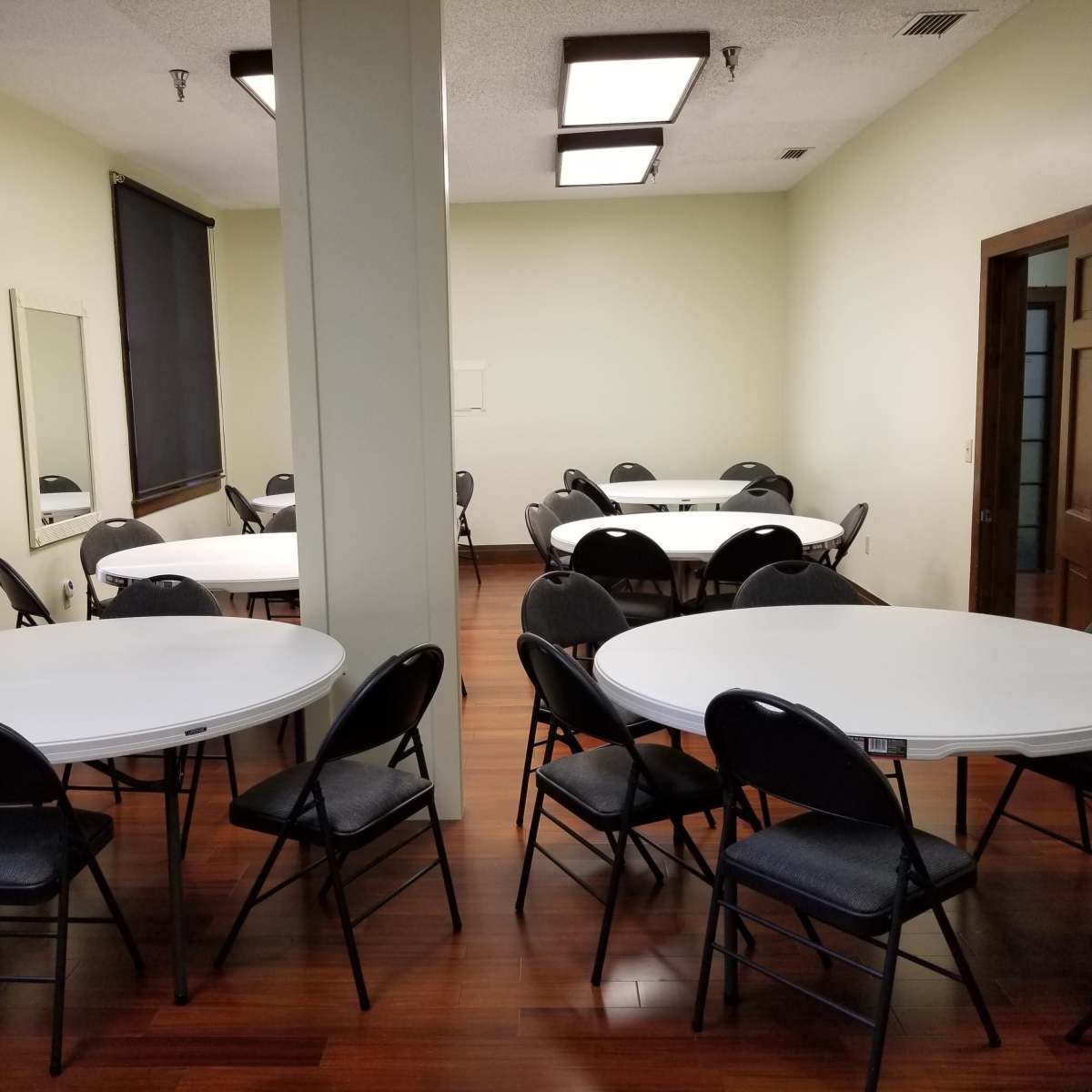 A room with multiple round tables and black chairs arranged around them, featuring hardwood flooring and large windows.