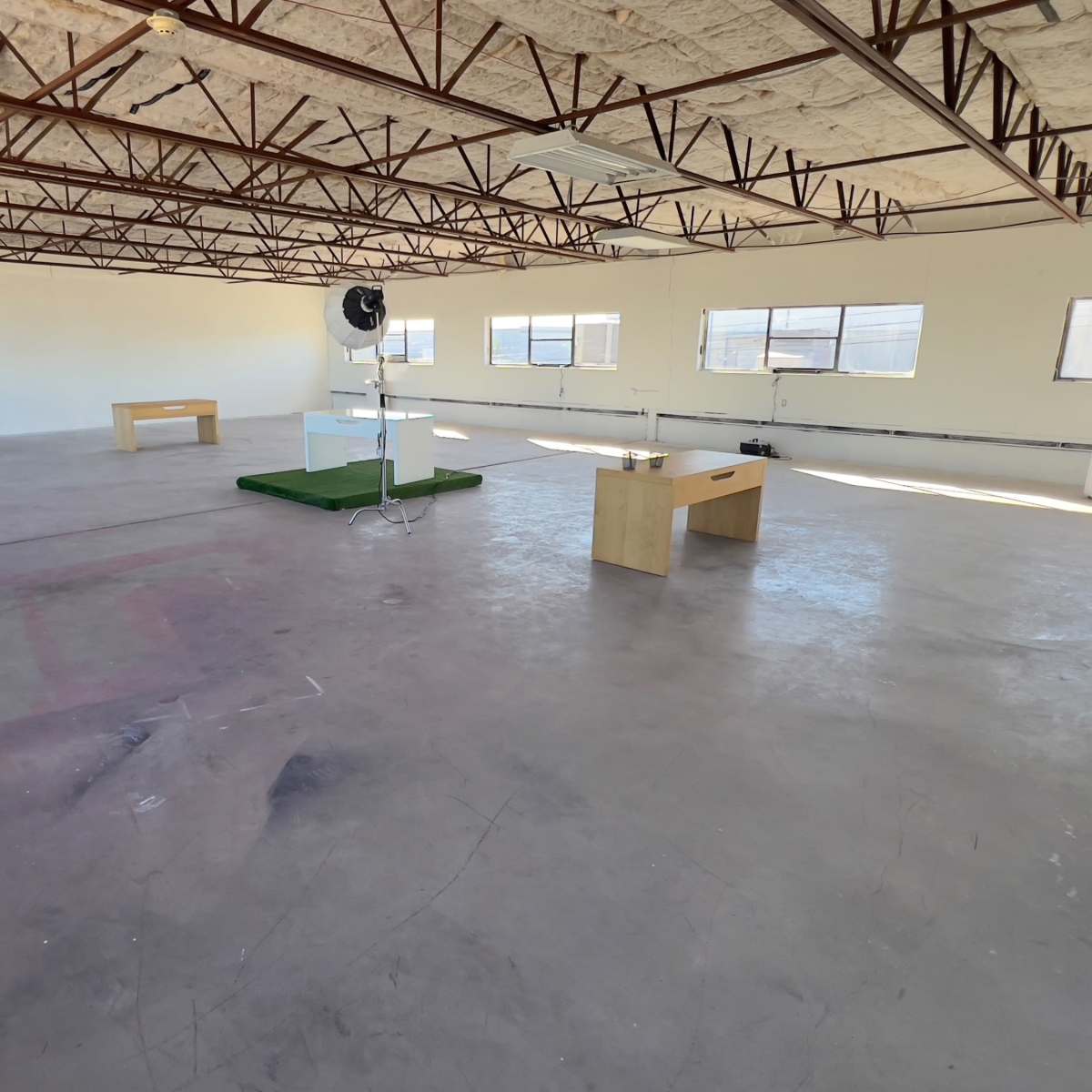 An empty room with a concrete floor, exposed metal beams, and minimal furniture including two wooden tables and a standing fan on a green mat.