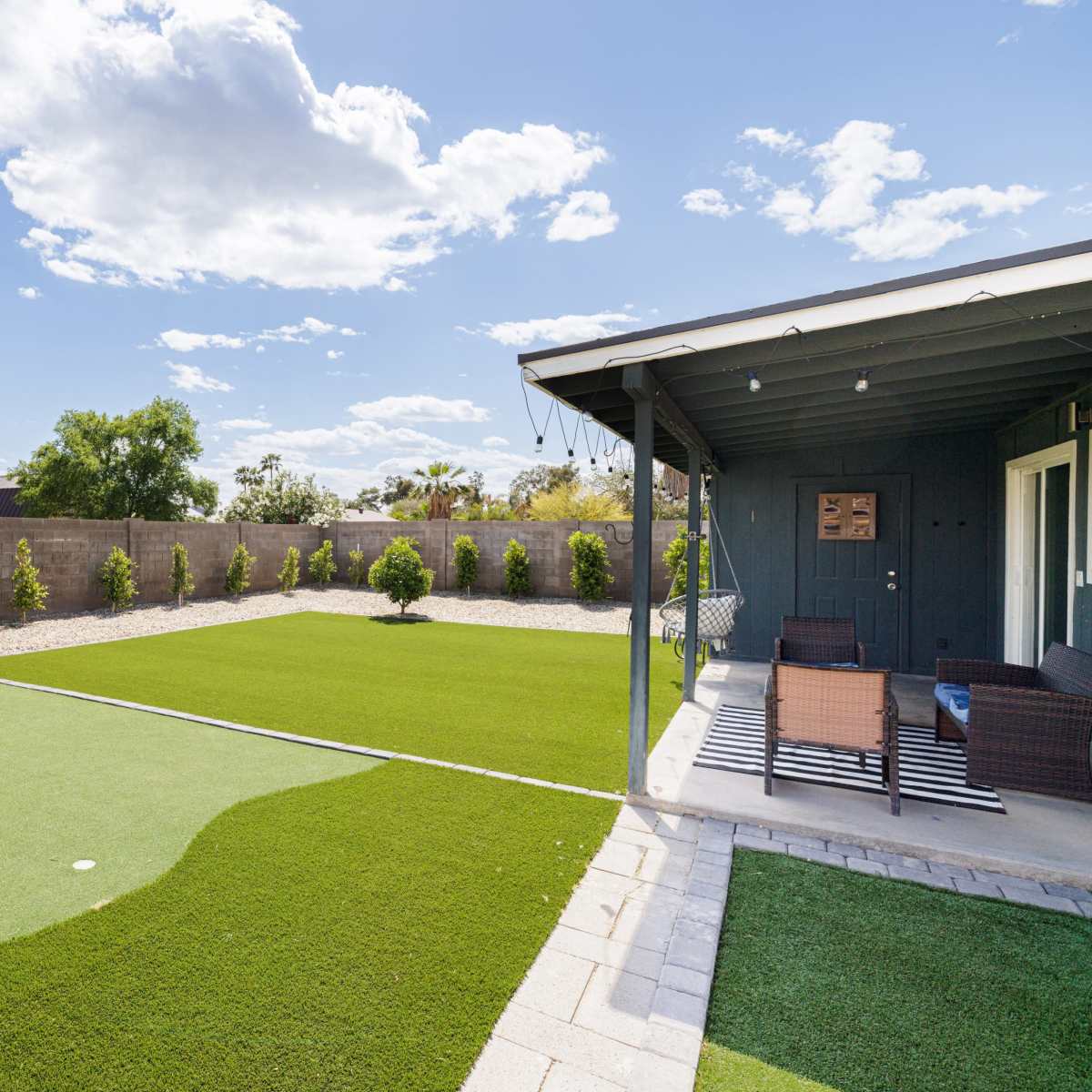 A landscaped backyard with artificial turf, bordered by low shrubs and a patio area with seating.