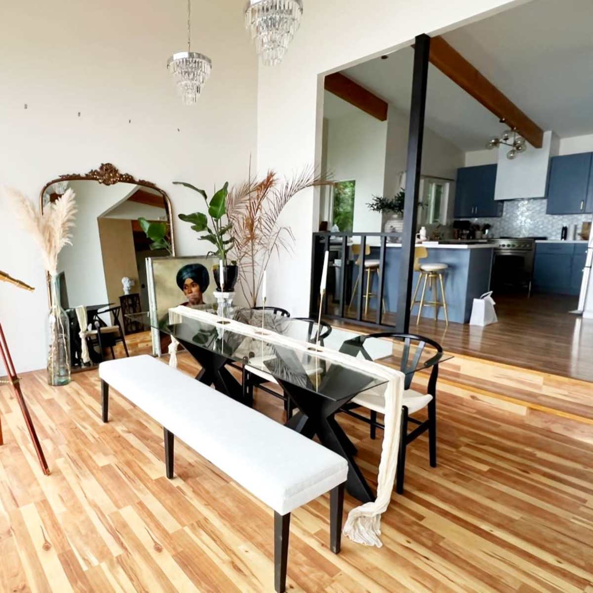 A modern dining area features a long table with a bench, surrounded by plants and decorative items, with an open kitchen visible in the background.
