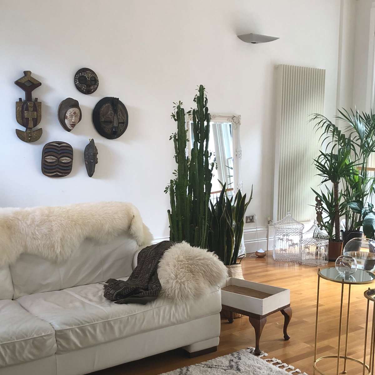 A bright living room with a white leather sofa, decorative wall masks, potted plants, a tall mirror, and a patterned area rug.