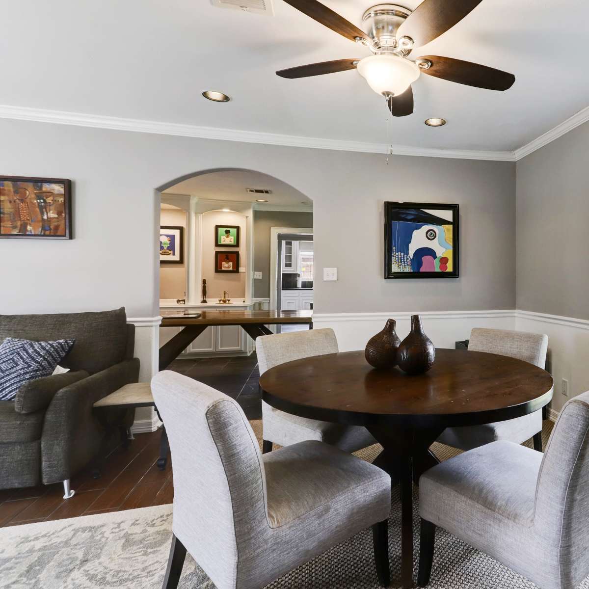 A round dining table is set with four upholstered chairs in a well-lit room featuring a ceiling fan and a window with a view of the outside.