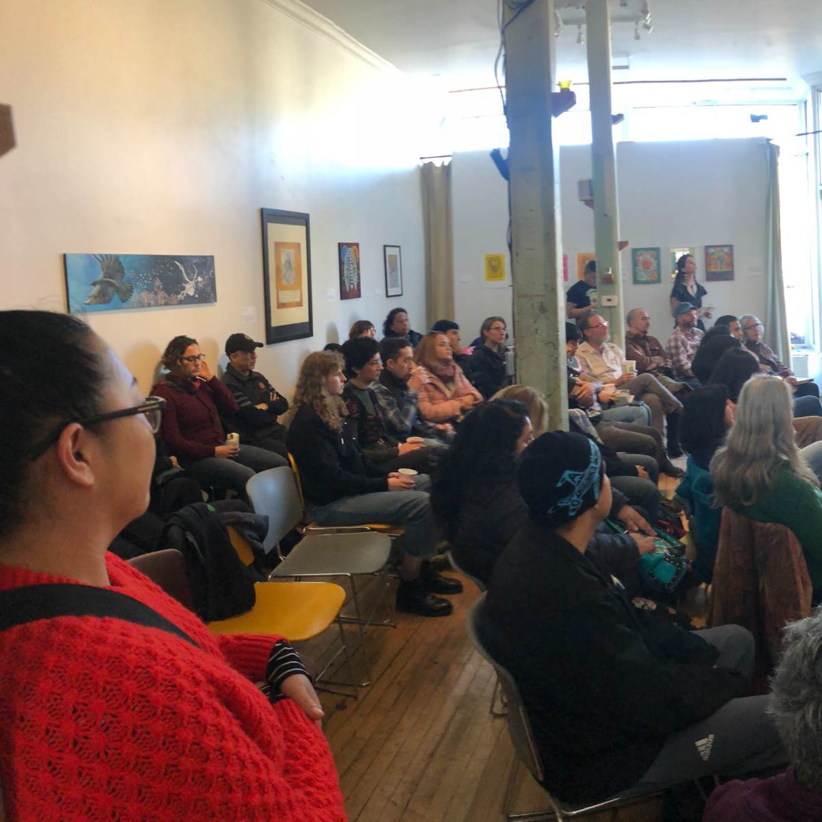 A speaker presents to an audience seated in a well-lit room with art on the walls.