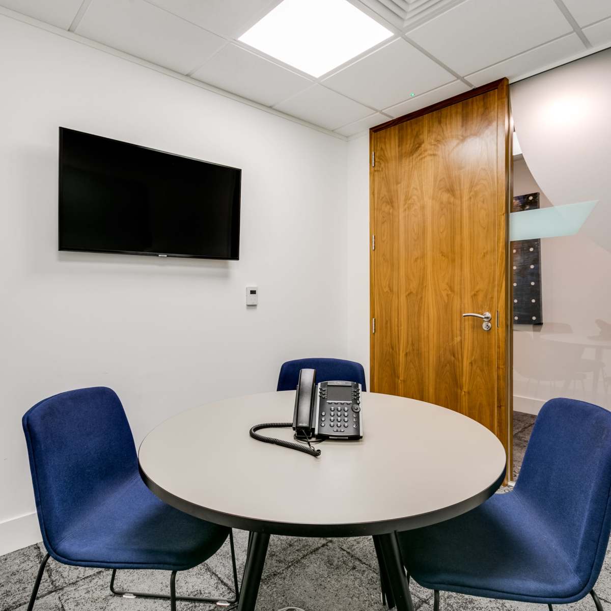 A small conference room features a round table with four blue chairs, a telephone, a wall-mounted TV, and a wooden door leading to another space.