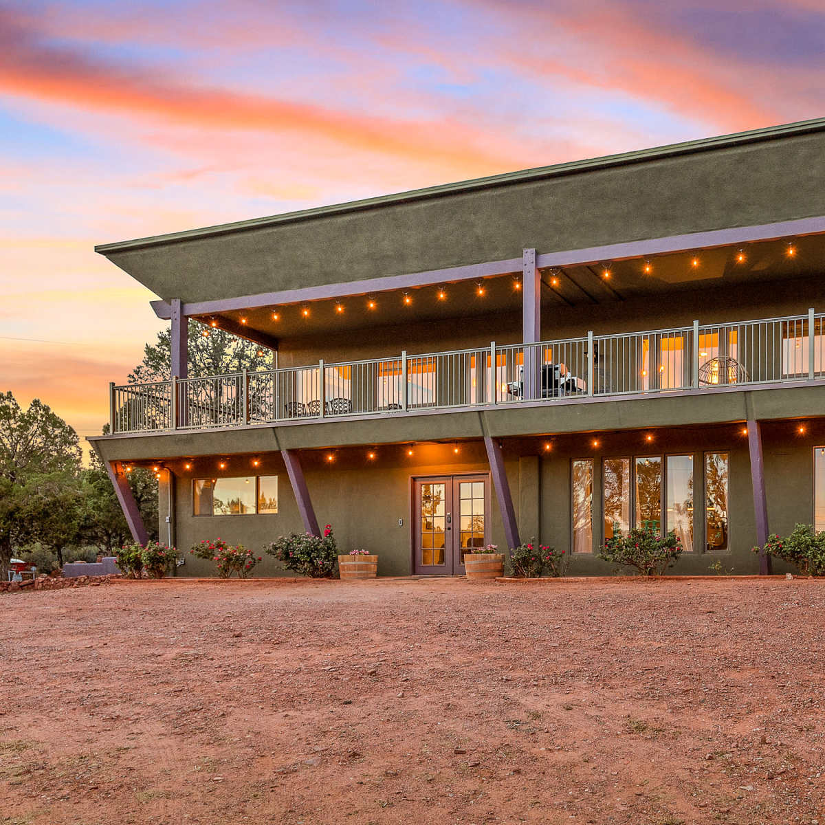 A modern two-story green house with a balcony, surrounded by gravel and landscaped plants, against a colorful sunset sky.