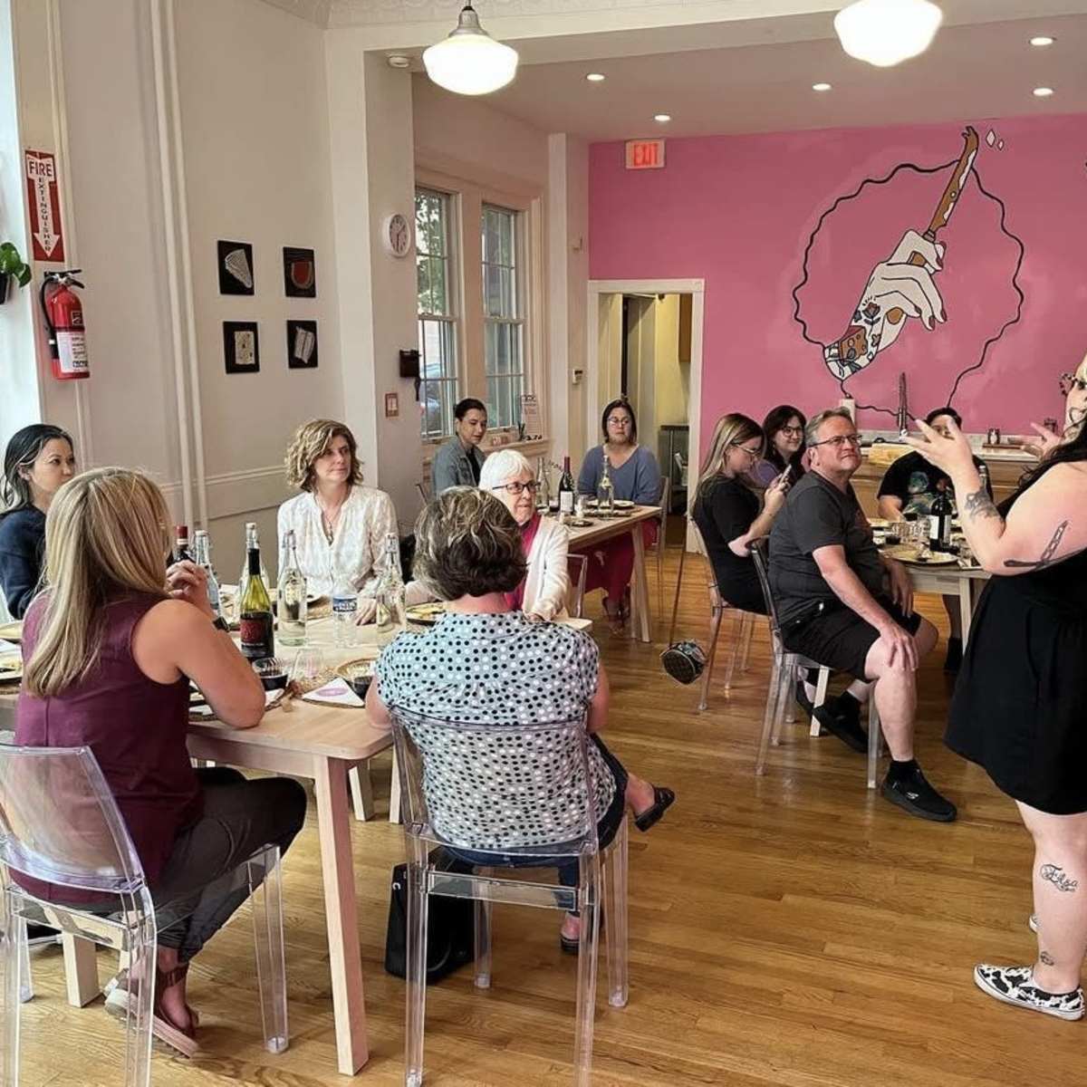A group of people sits at tables in a café while a woman stands and speaks in front of a painted mural.