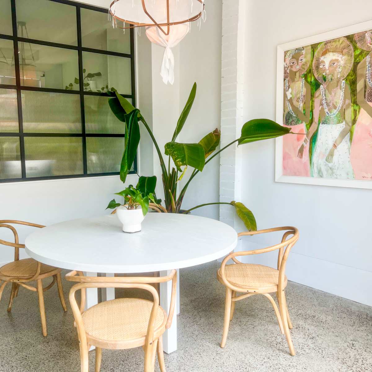 A round white table with four woven chairs is positioned in a bright room featuring a large window and a potted plant.