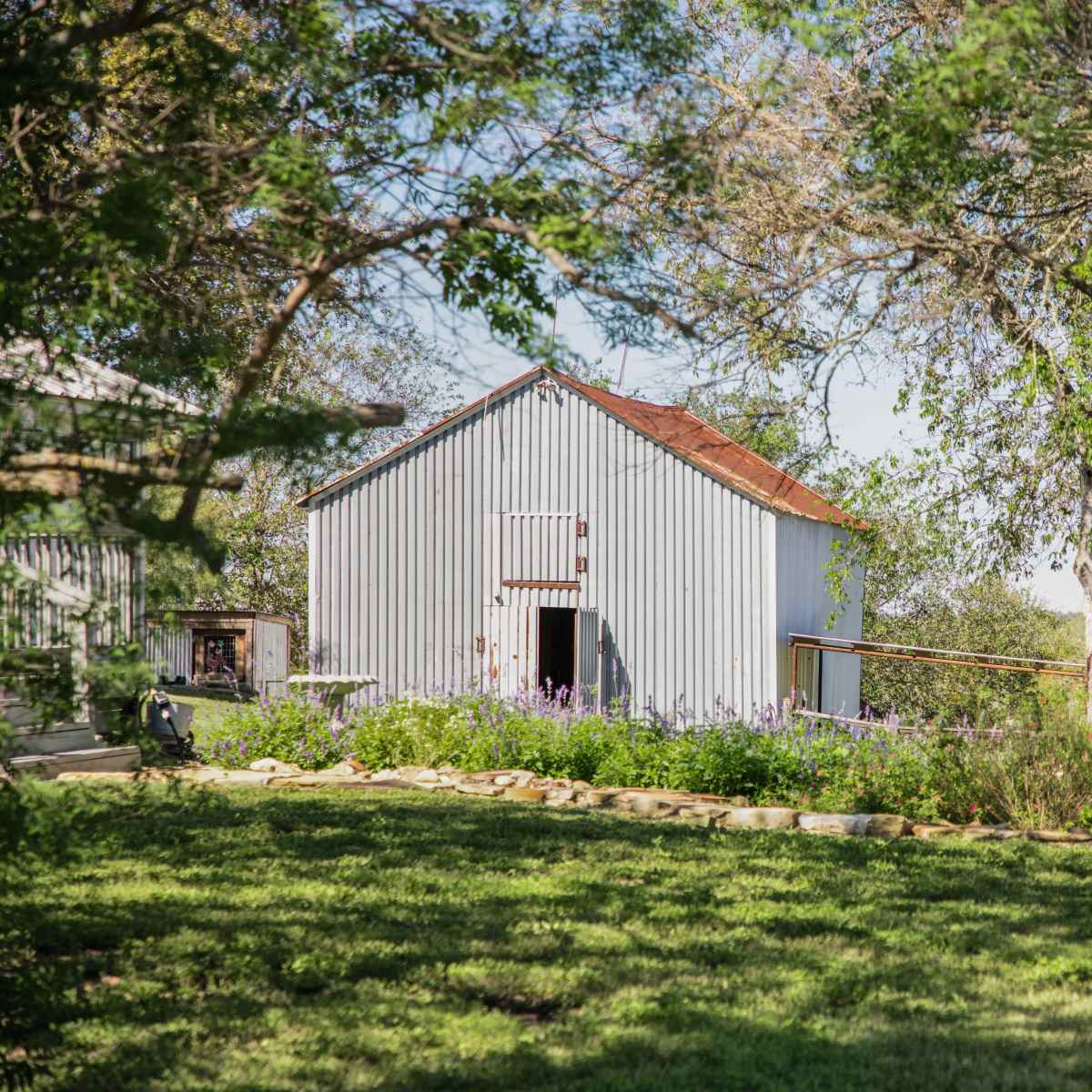 A white barn stands surrounded by trees and flowering plants in a clear outdoor setting.