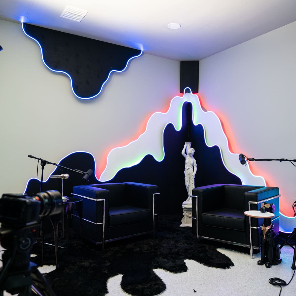 A modern recording setup with two black chairs, a decorative wall with colorful neon lights, and multiple cameras positioned around the space.