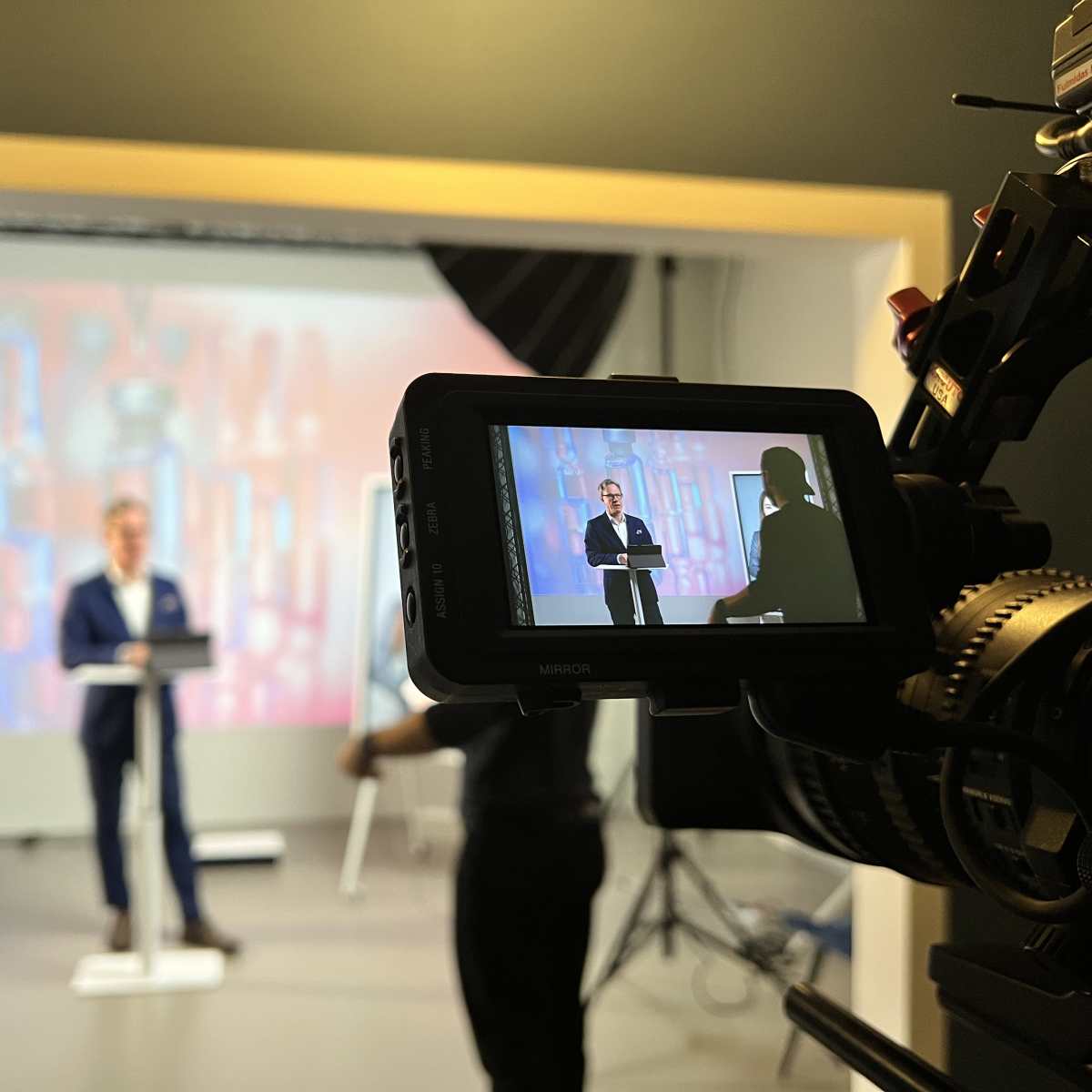 A speaker in a blue suit addresses an audience while standing behind a podium in a studio setting.