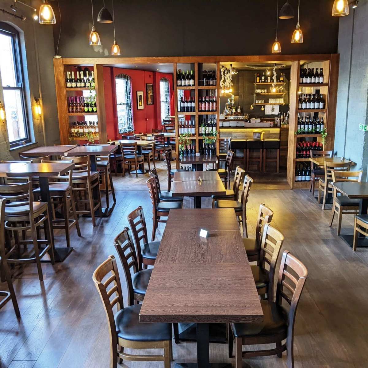 A spacious restaurant interior with wooden tables and chairs arranged around a central bar, flanked by shelves filled with bottles.